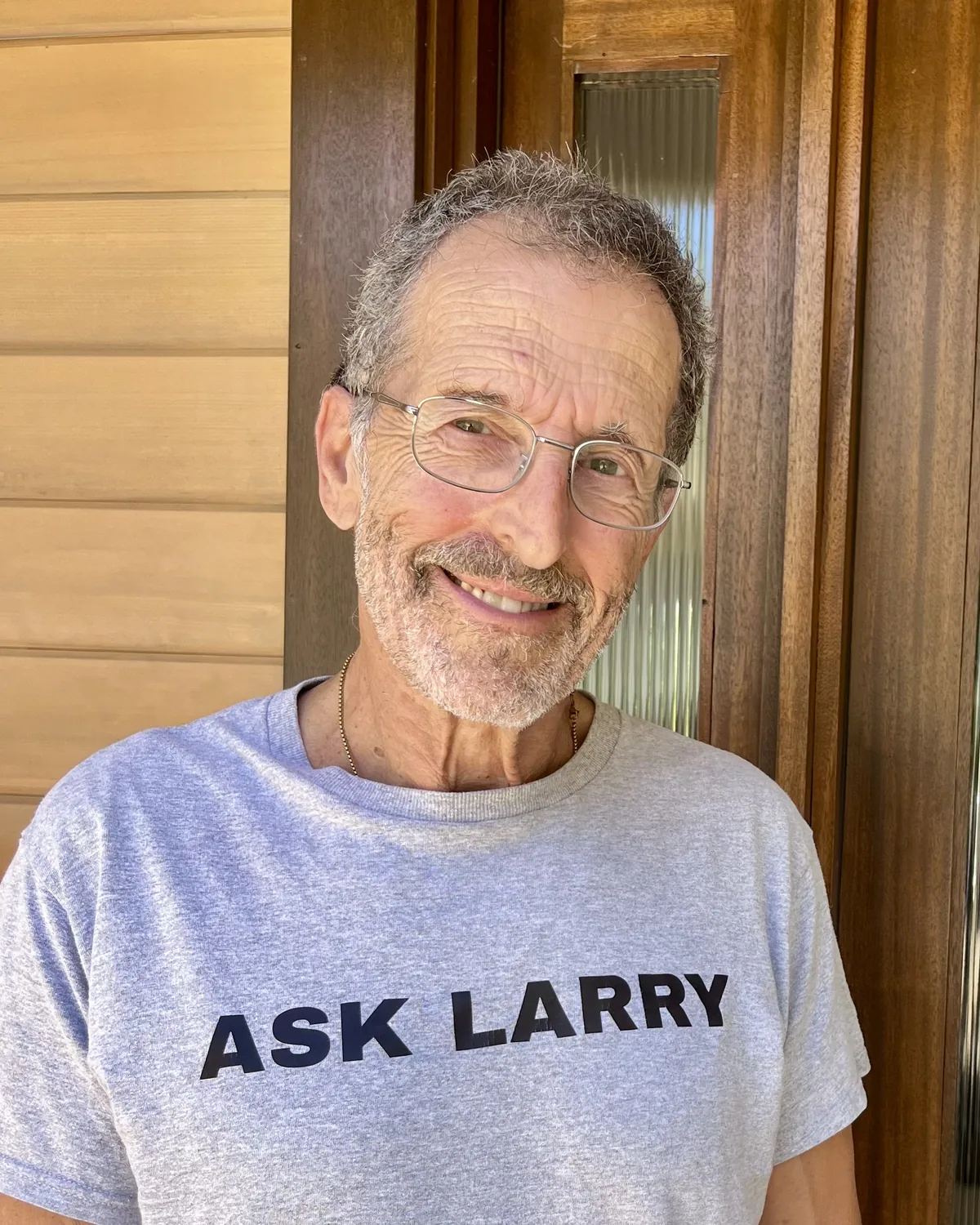 Larry Steingold, smiling, on his front porch in a grey ASK LARRY t-shirt.